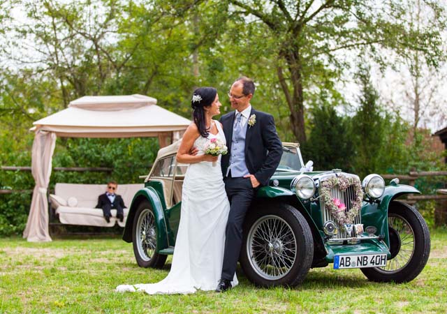 Hochzeitspaar angelehnt an grünem Oldtimer vor grüner, romantischer Natur Kulisse